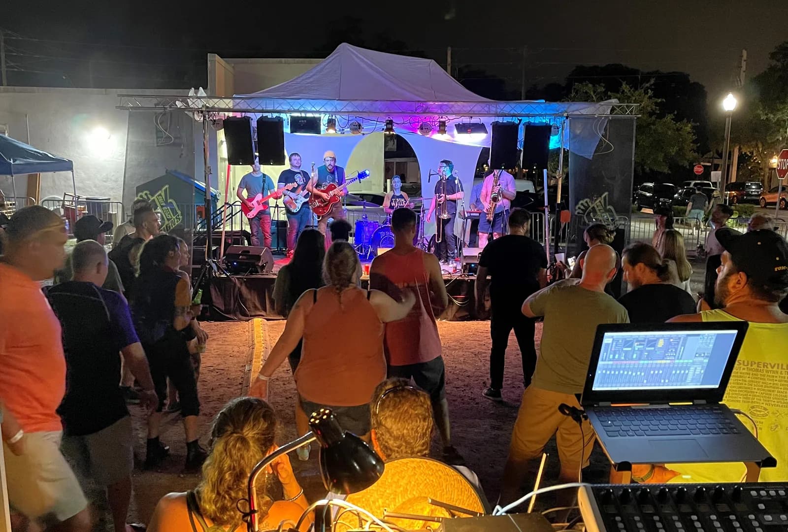 Front of house position at an outdoor evening event, with a mixing console and laptop looking out toward a lit stage and the crowd