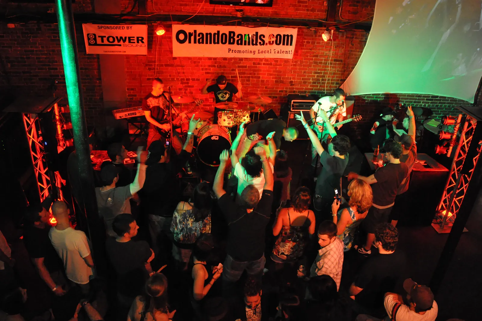 A packed brick-walled venue with a band performing under red and green stage lighting and the crowd close to the stage, beneath an OrlandoBands.com banner