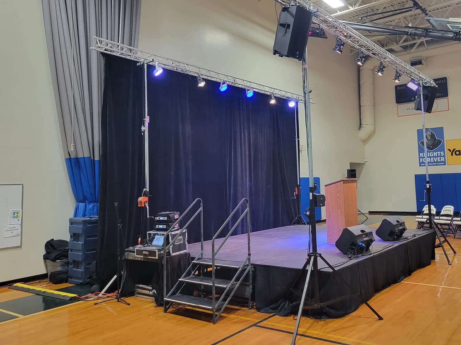 A CFAV setup in a school gymnasium: a low stage deck and curtain backdrop under a lighting truss of blue LED fixtures, a main speaker on a stand at each side of the stage, wedge monitors along the deck edge, a podium, and an amp rack and small console on a road case at the side