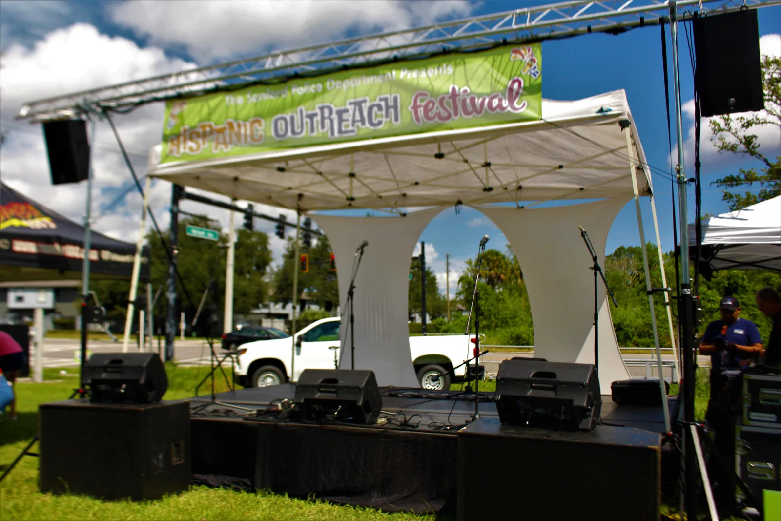 Outdoor festival stage set before the event, with vocal microphones, stage monitors, and speakers in place beneath a banner