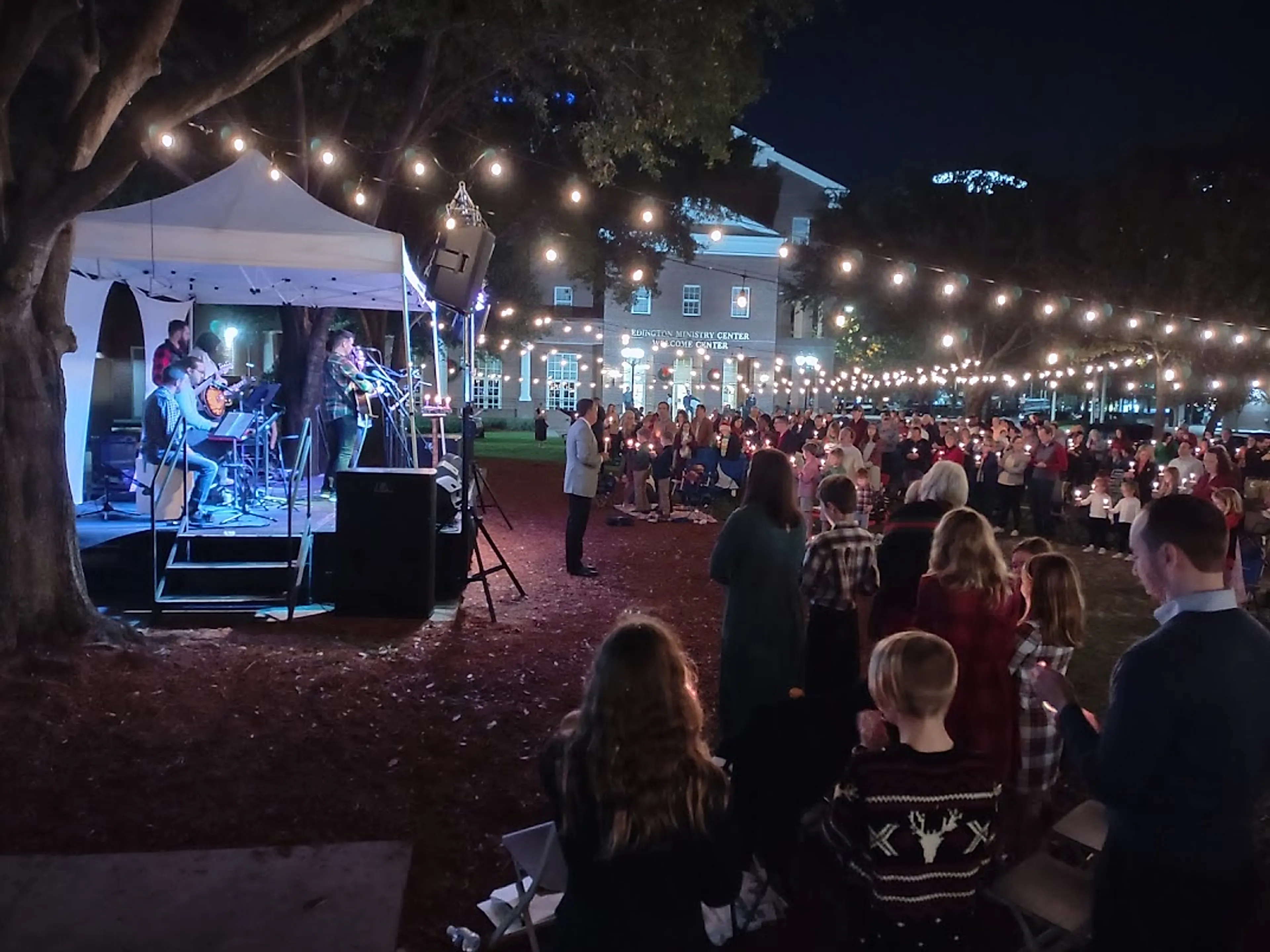 An evening outdoor service produced by CFAV, with a band playing on a covered stage under strings of lights and the congregation holding candles