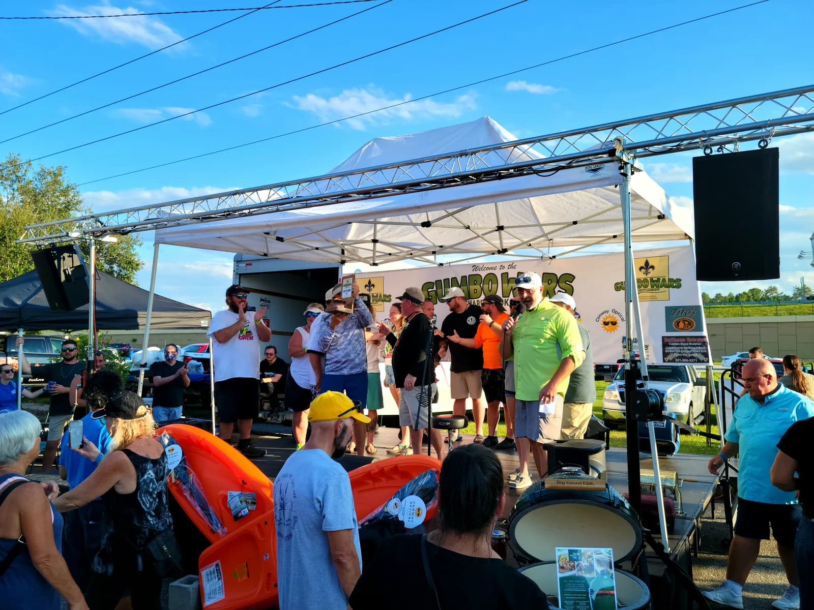Daytime outdoor festival stage under a canopy with speakers on truss, a presenter on a microphone, and a crowd gathered in front