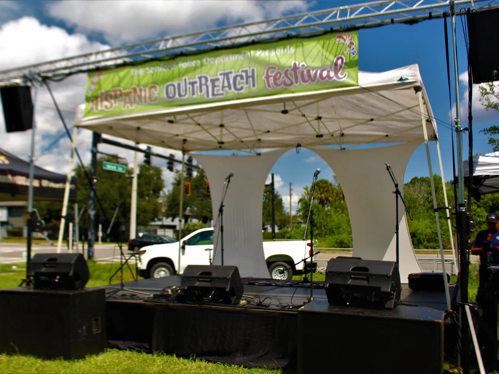 Outdoor festival stage with a banner overhead, stage monitors along the deck, and speakers on truss on either side