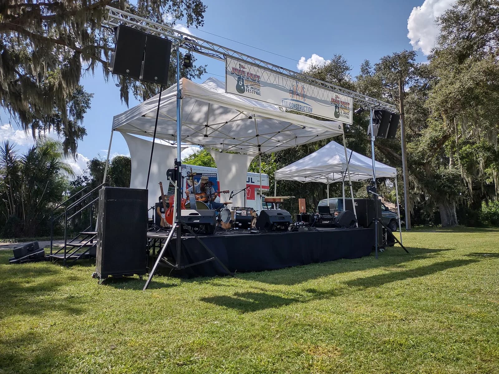 Outdoor festival stage with a white canopy roof, speakers on truss, and instruments set up under oak trees