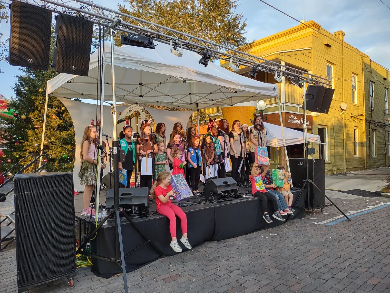 Children singing on an outdoor street stage in the late afternoon, with speakers and stage lighting on truss