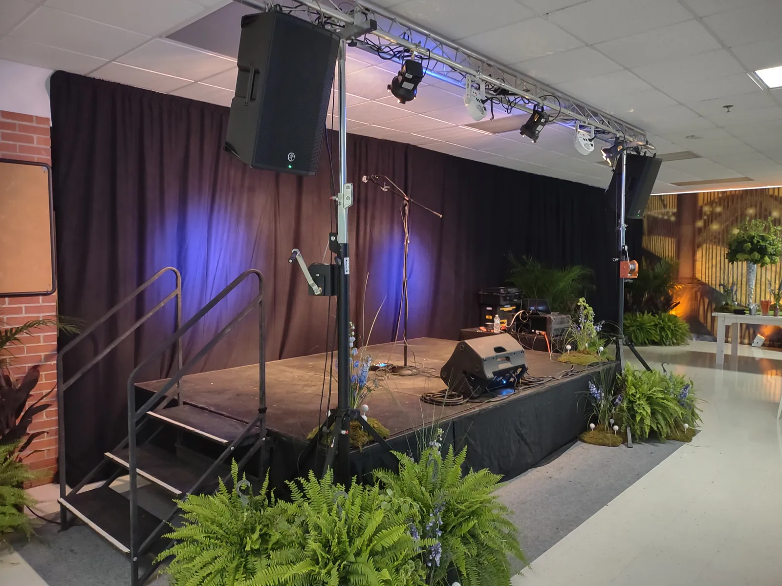 Indoor stage dressed with plants and lit in blue and purple, with microphones and stage monitors set for a ceremony