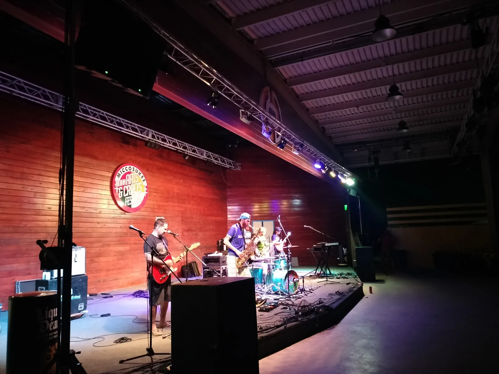 Band performing on an indoor stage against a wood wall, lit by stage lighting mounted on truss