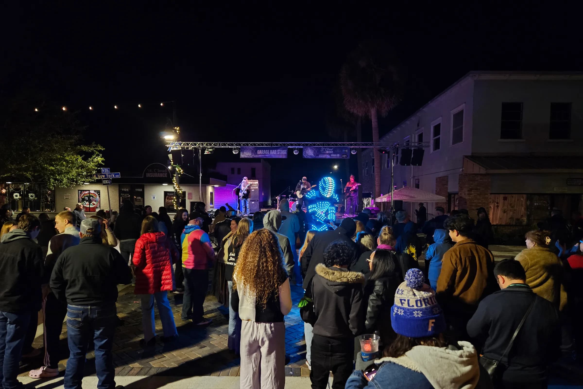 Night crowd watching a band perform on an outdoor street stage lit in blue and purple