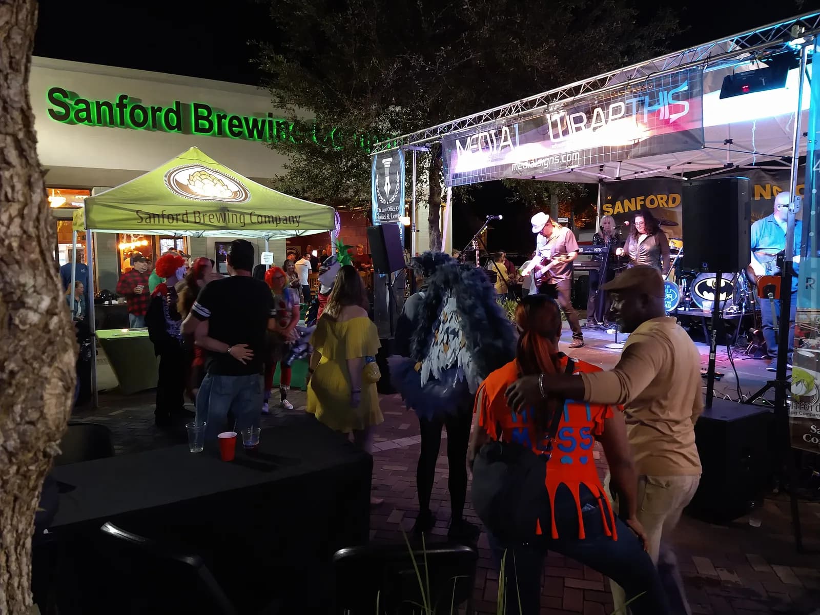 Band performing outdoors at night beside a brewery, with stage lighting on truss and a crowd in costume