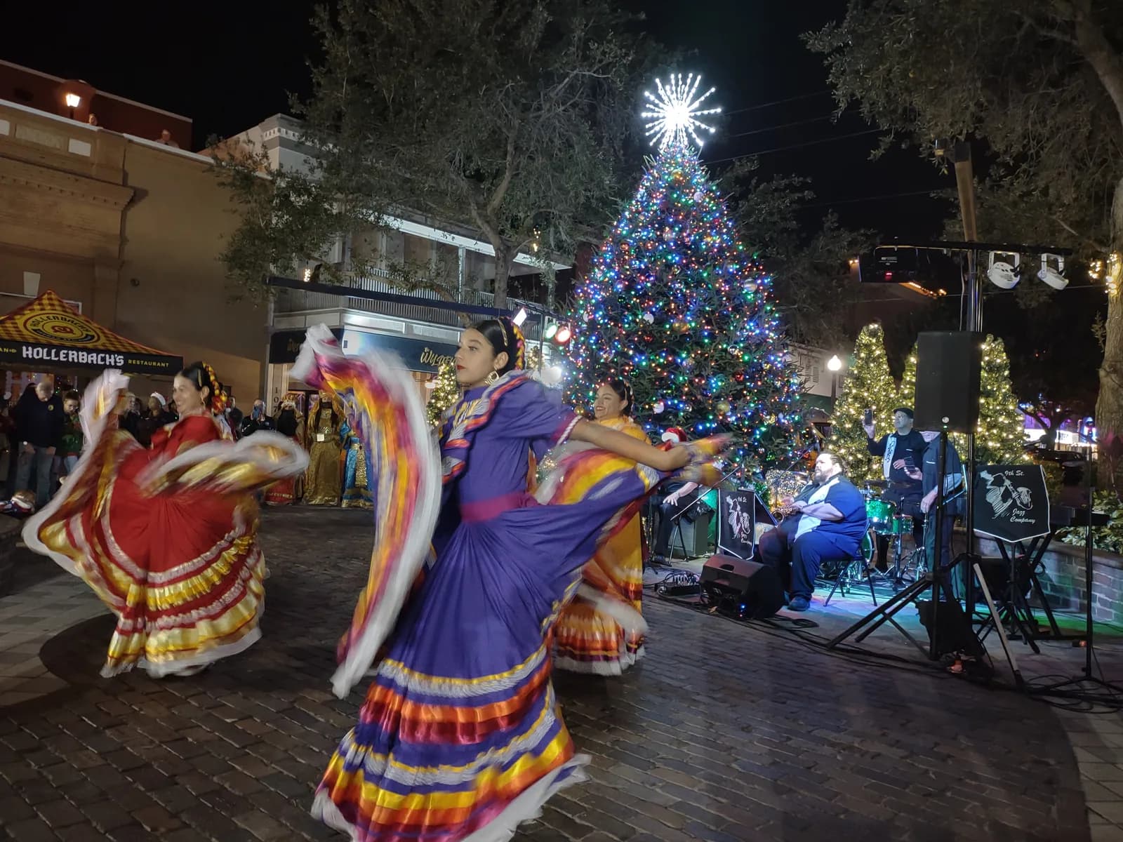 Dancers in traditional dress performing at night beside a lit holiday tree, with a stage and speakers behind them