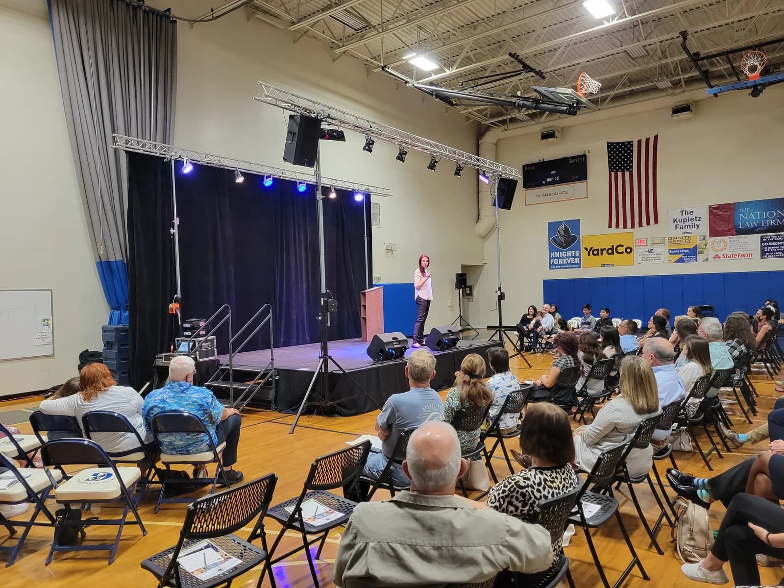 Speaker presenting on a stage set up inside a school gym, with lighting on truss and an audience in folding chairs