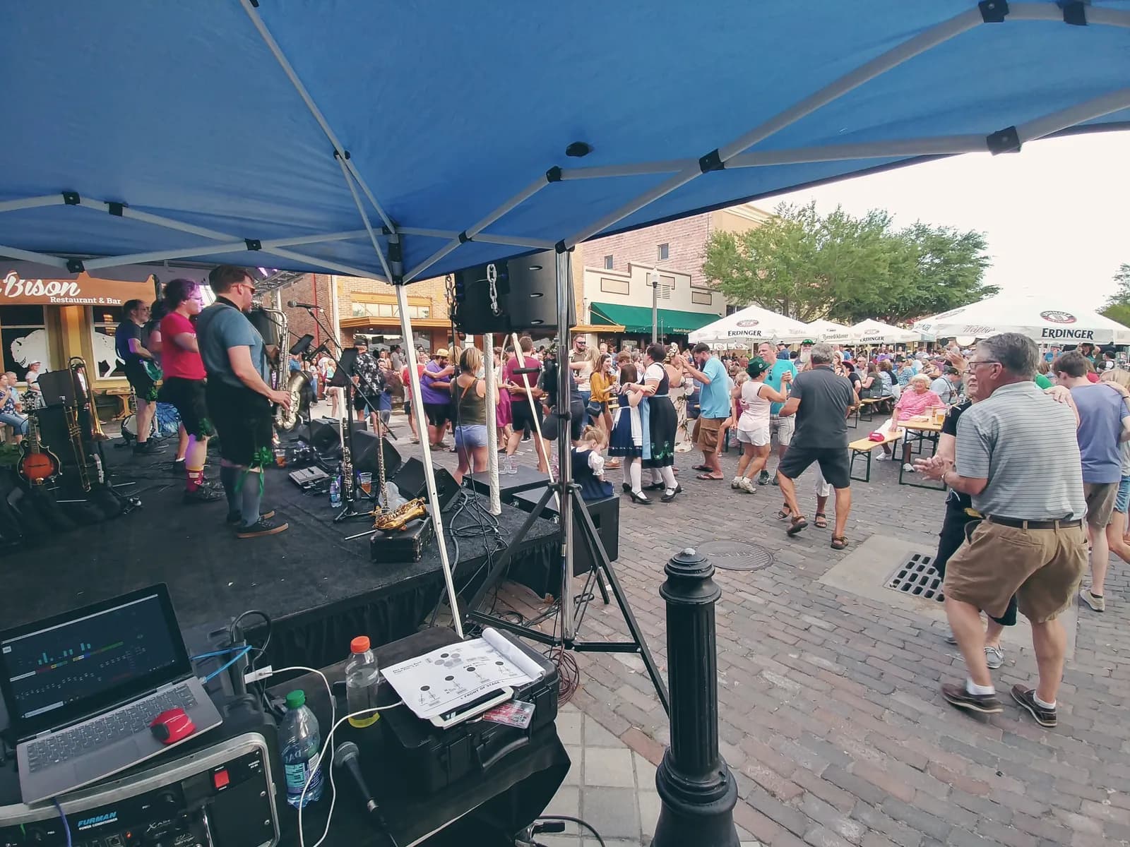 Mixing console at the front of house position under a tent, looking out at a band and a crowd on a brick street