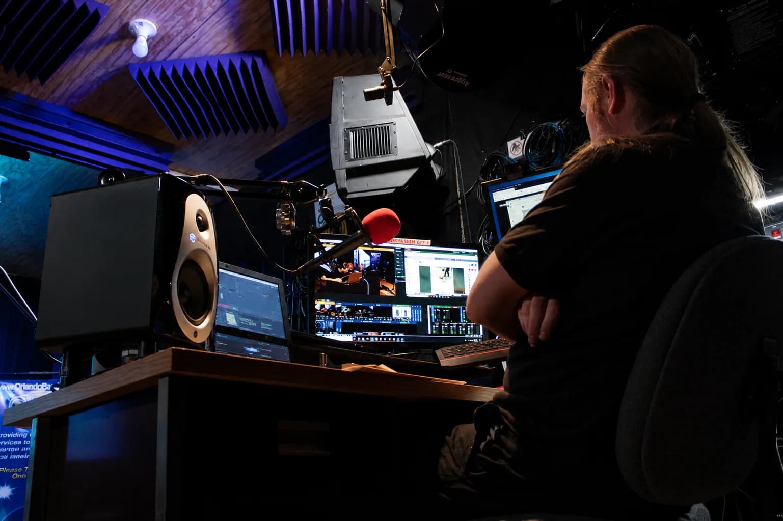 The production position in the CFAV studio: a desk of screens, a studio monitor speaker and a microphone on a boom arm, with someone seated at it, below the room's acoustic panelling